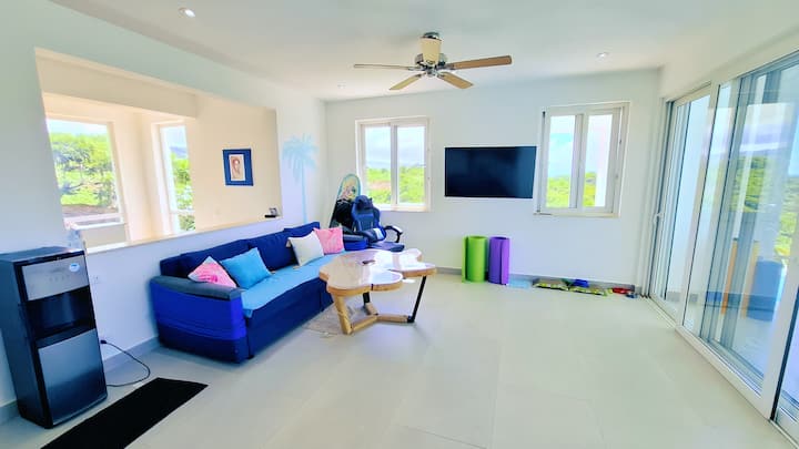 Sala de estar luminosa y espaciosa con vistas al mar y a la piscina infinita. Decoración tropical, asientos cómodos, TV inteligente, dispensador de agua y una mesa para descansos, lectura o trabajo a distancia.