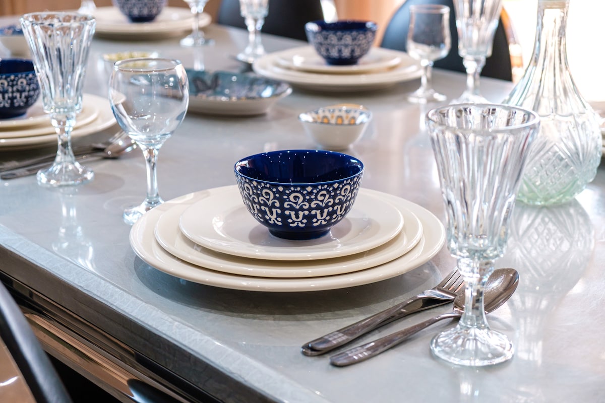 A dining table is set with neatly arranged dinnerware, including a blue decorative bowl placed centrally on white plates. Crystal glasses and cutlery are positioned uniformly, creating a welcoming atmosphere for mealtime. Natural light enhances the table setting, illuminating the polished surface.