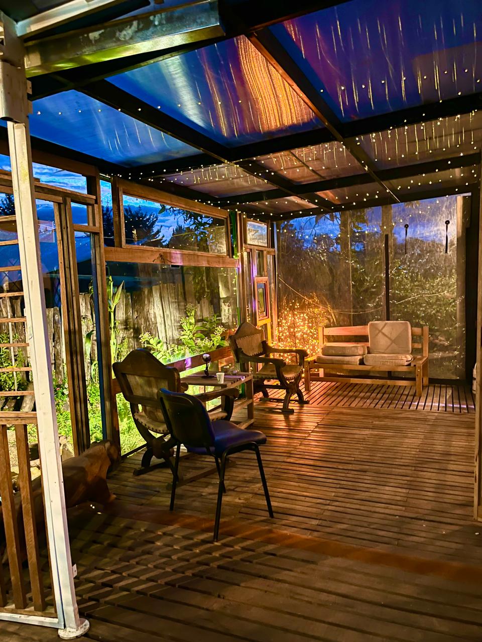 A covered terrace area featuring wooden flooring is illuminated by soft lighting. Several seating options are visible, including two chairs and a bench. Green plants surround the space, creating a tranquil ambiance. Large transparent panels provide views of the outdoor surroundings.