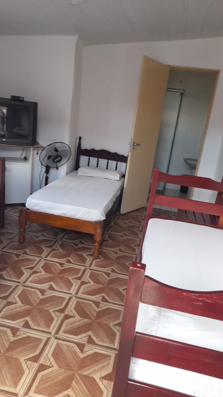 A simple room layout includes two twin beds positioned against the wall, complemented by a wooden nightstand. A television sits on a small table near the door, and a standing fan is visible in the corner. The floor features a geometric patterned design.