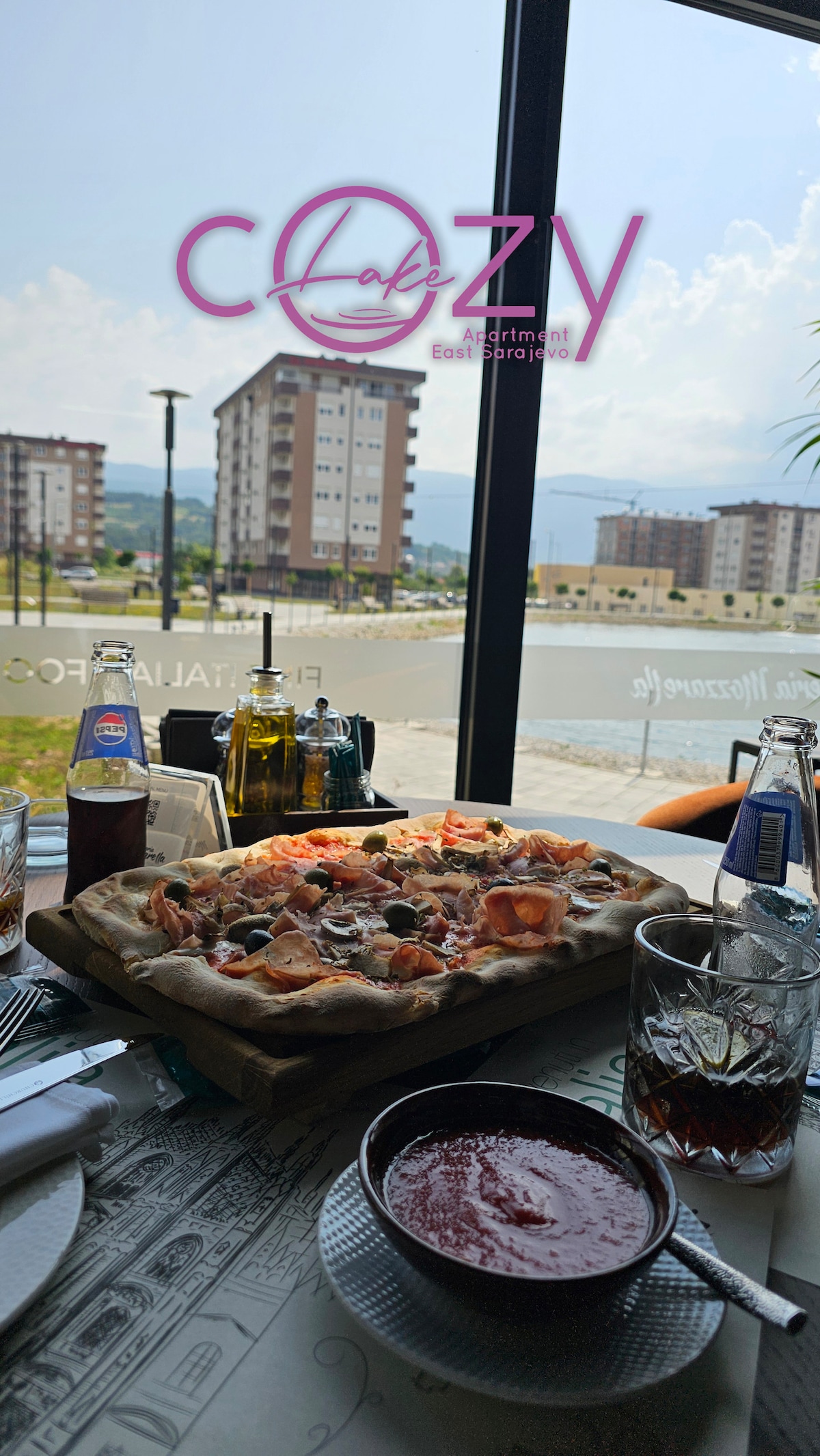 A dining table is set with a freshly made pizza topped with various ingredients. Nearby, drinks are displayed in glasses and bottles. A large window offers a view of buildings and a clear sky, providing natural light to the inviting dining space.