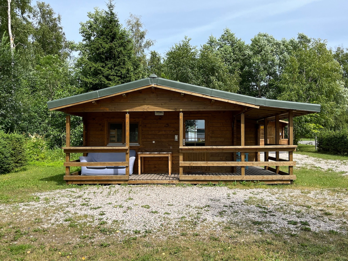 The cabin's exterior features a wooden facade with a green roof, surrounded by lush greenery. A spacious porch with seating is visible, offering a comfortable spot to enjoy the natural setting. Gravel and grass cover the ground, enhancing the rustic charm.