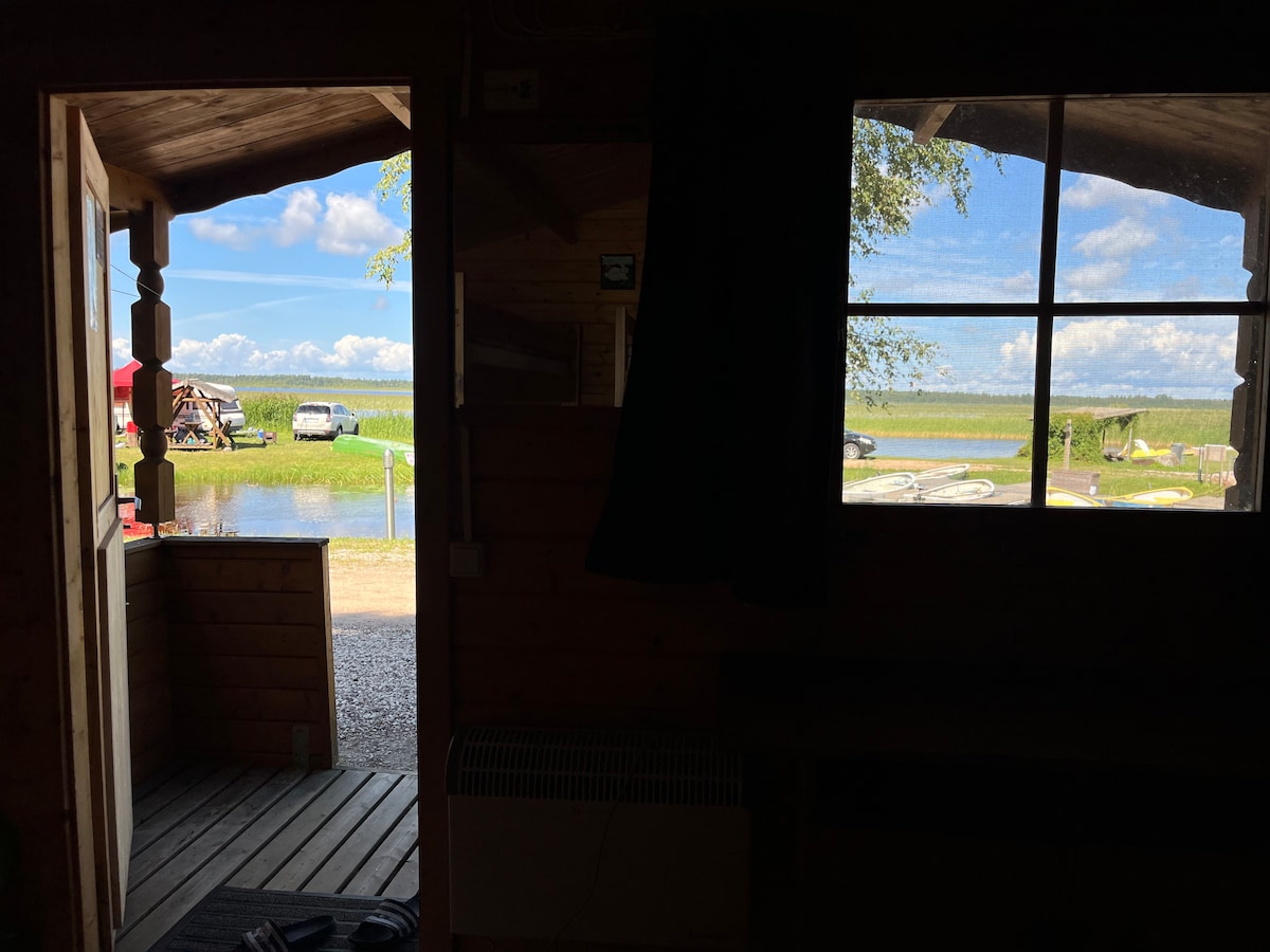 A view is captured from inside the cabin, featuring an open door that leads to a deck area. Scenic outdoor space is visible, showcasing a calm lake and lush greenery under a bright sky. Natural light enters through the door and a nearby window.