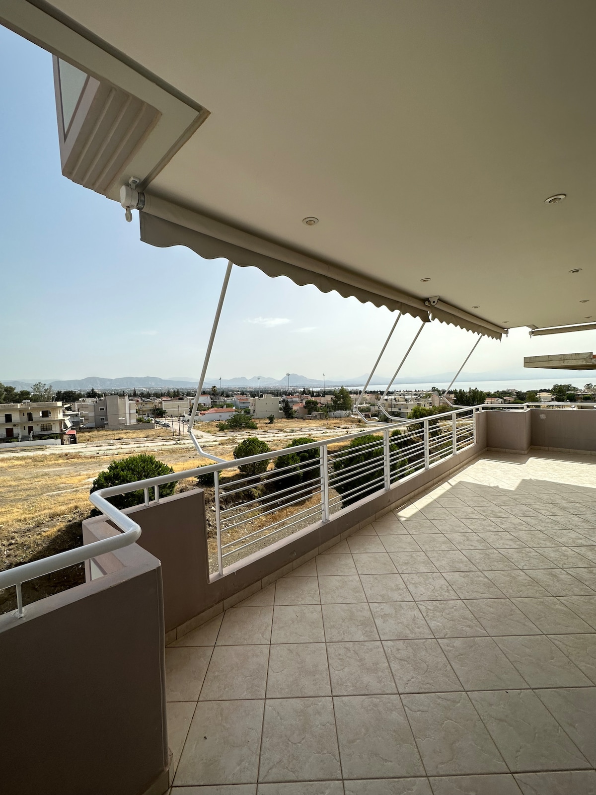 A spacious balcony offers a panoramic view of the surrounding landscape, featuring rolling hills and a clear sky. The tiled floor extends outward, with protective awnings partially shading the area. An unobstructed sightline enhances the inviting atmosphere for outdoor relaxation.