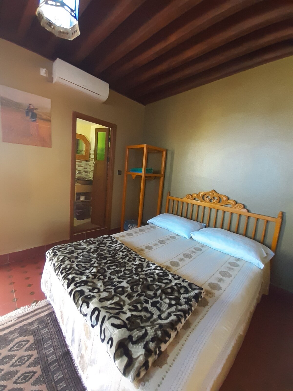 A cozy sleeping area features a queensize bed adorned with a patterned blanket. Light pours in from a nearby window, and a wooden cabinet stands against the wall. A doorway leads to a visible private bathroom, enhancing the overall comfort of the room.