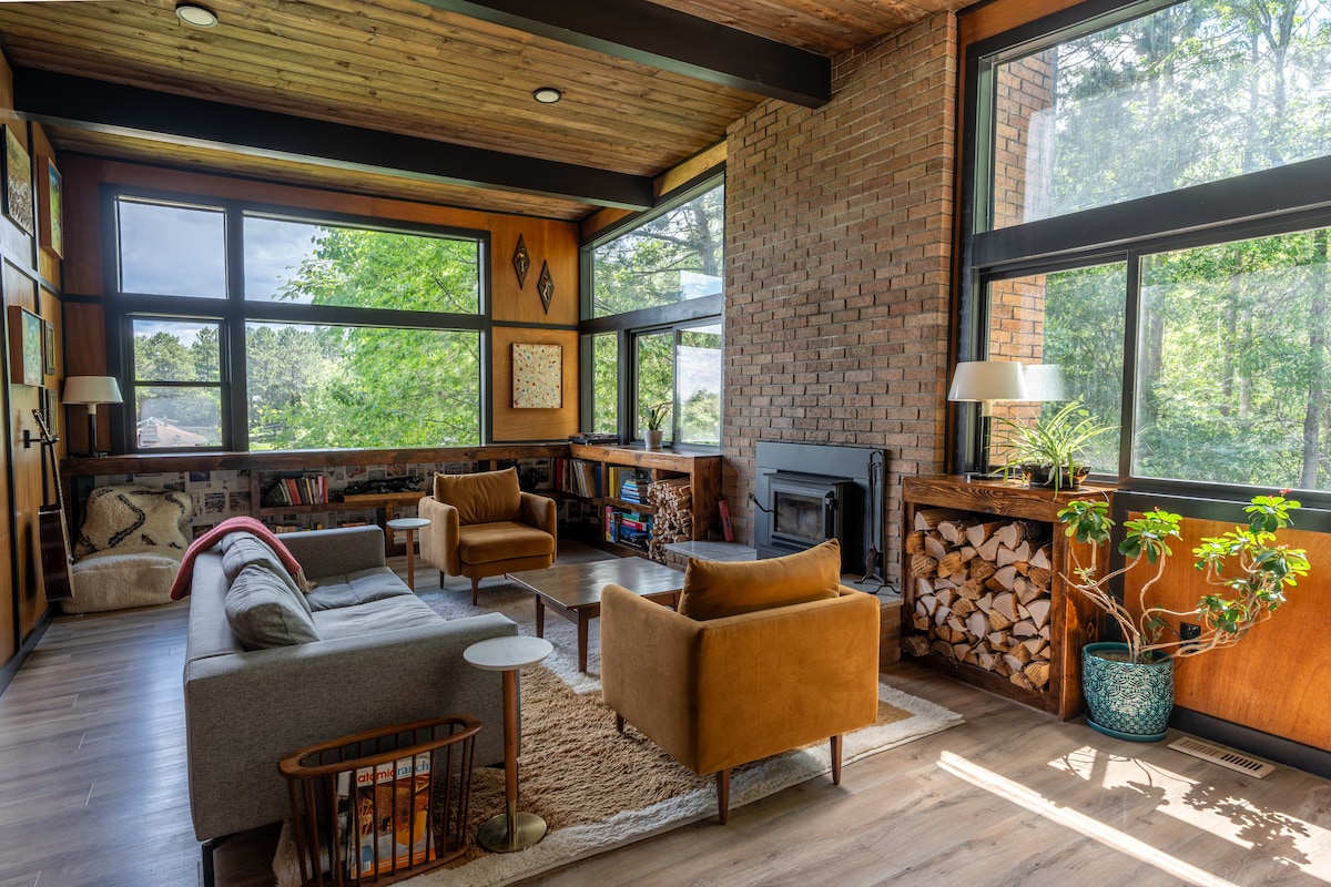 A cozy living area features large windows allowing natural light to fill the space, showcasing views of surrounding greenery. A gray sofa and two brown armchairs are arranged around a central coffee table. A wood-burning fireplace is flanked by stacked firewood, with a decorative plant adding a touch of nature.