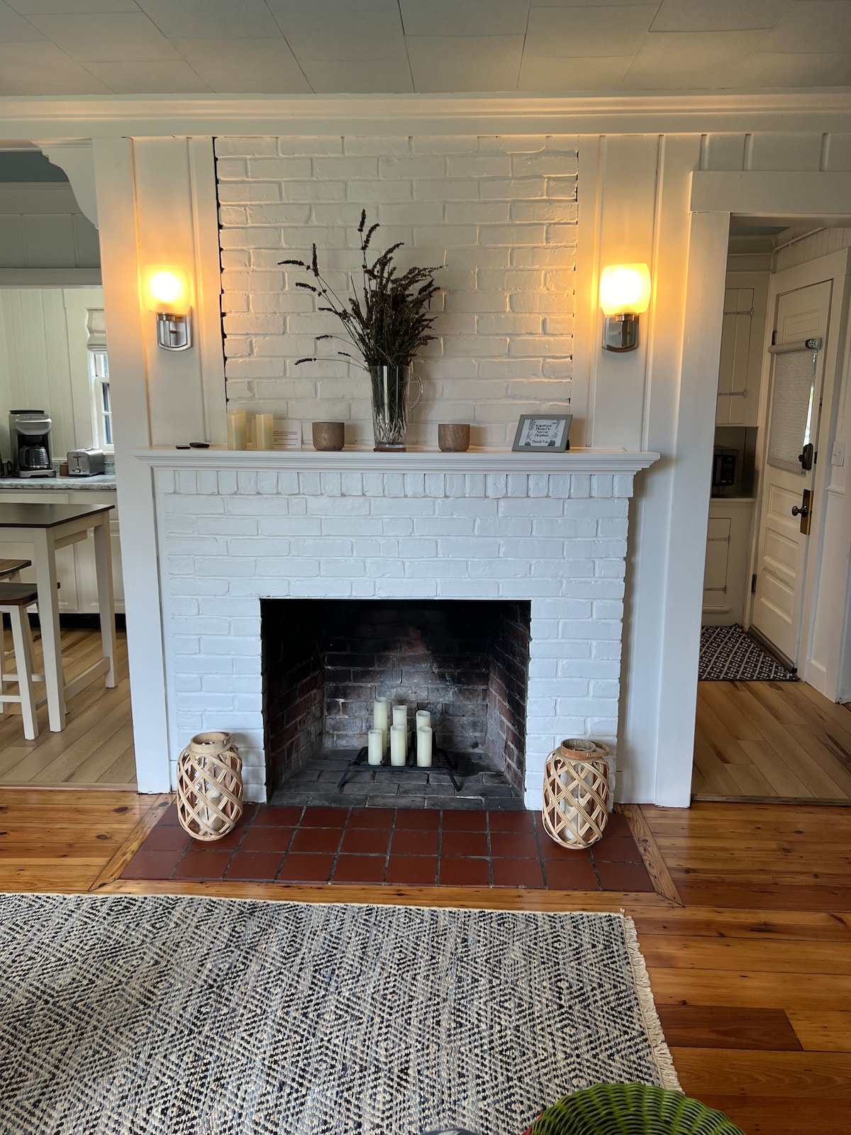 A charming fireplace, framed by white brick, creates a focal point in the living area. Neutral tones are complemented by wooden accents. A textured area rug lies on the floor, while a decorative vase with dried plants adorns the mantel, enhancing the cozy ambiance.