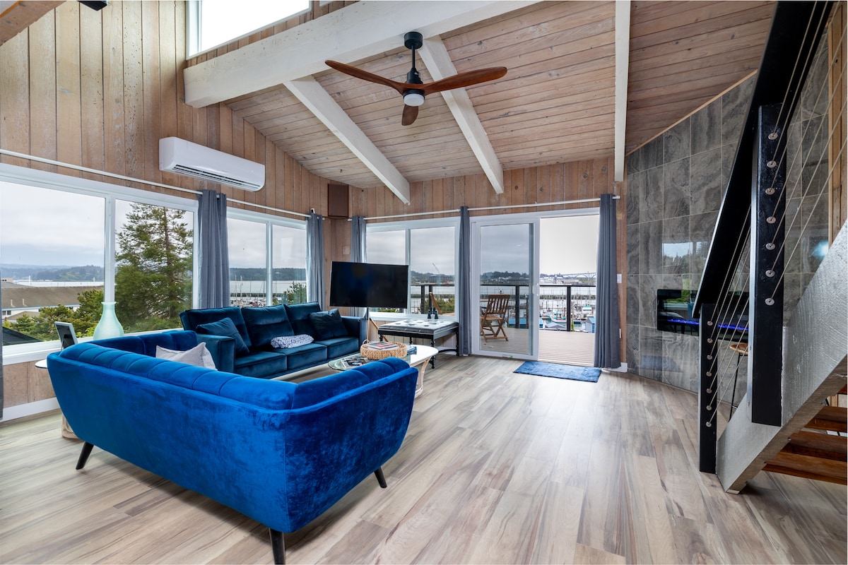 A bright living area features large windows providing views of the harbor. A blue sofa is positioned for comfort, with a modern television nearby. Light wood beams enhance the openness, while a staircase adds an architectural element. A cozy rug and decorative accents complete the space.