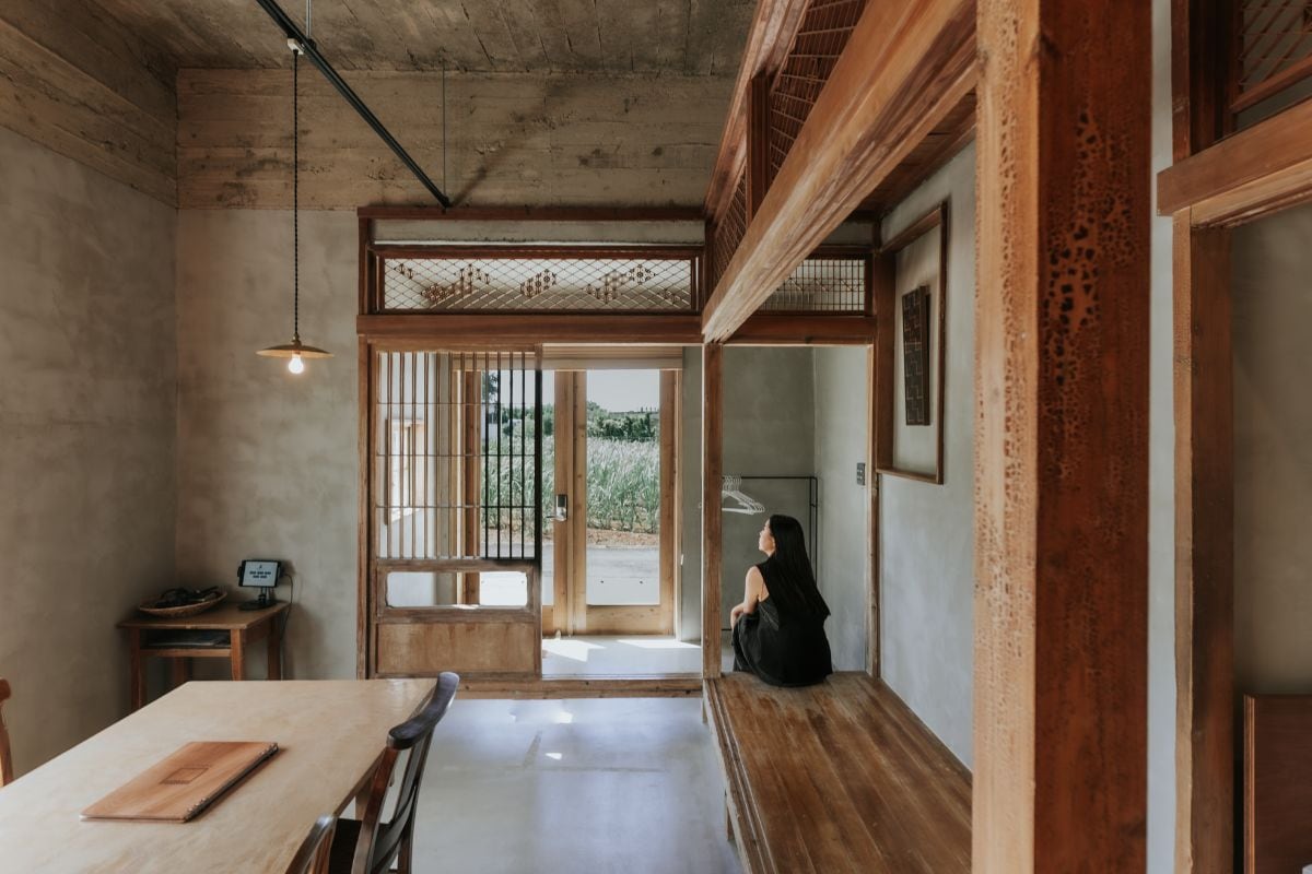 A spacious interior showcases traditional wooden architecture with an open layout. Natural light filters through sliding doors that lead to the outside area. A dining table is positioned nearby, with a cozy seating area featuring a wooden floor adjacent to the entrance.