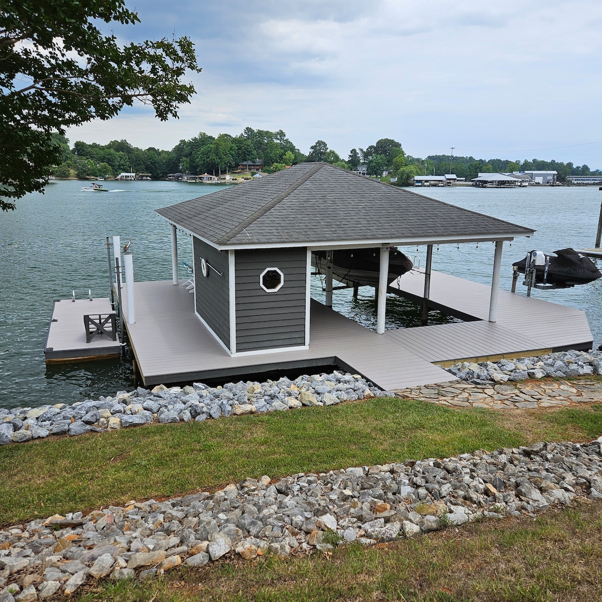 A spacious dock features a covered lounging area with a circular window, surrounded by calm waters. A wooden walkway extends from the shore, lined with smooth stones. Nearby boats are visible, contributing to a serene lakeside atmosphere.