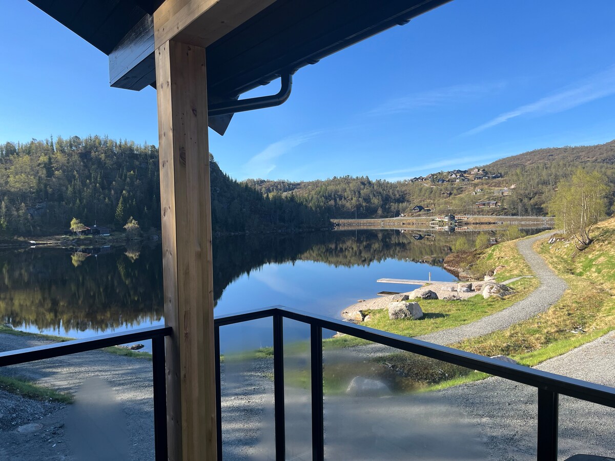 The image captures a serene lake view from a cabin terrace, framed by wooden beams. Reflections of the surrounding hills and trees are visible in the calm water. A gravel path leads along the lakeshore, inviting outdoor exploration.
