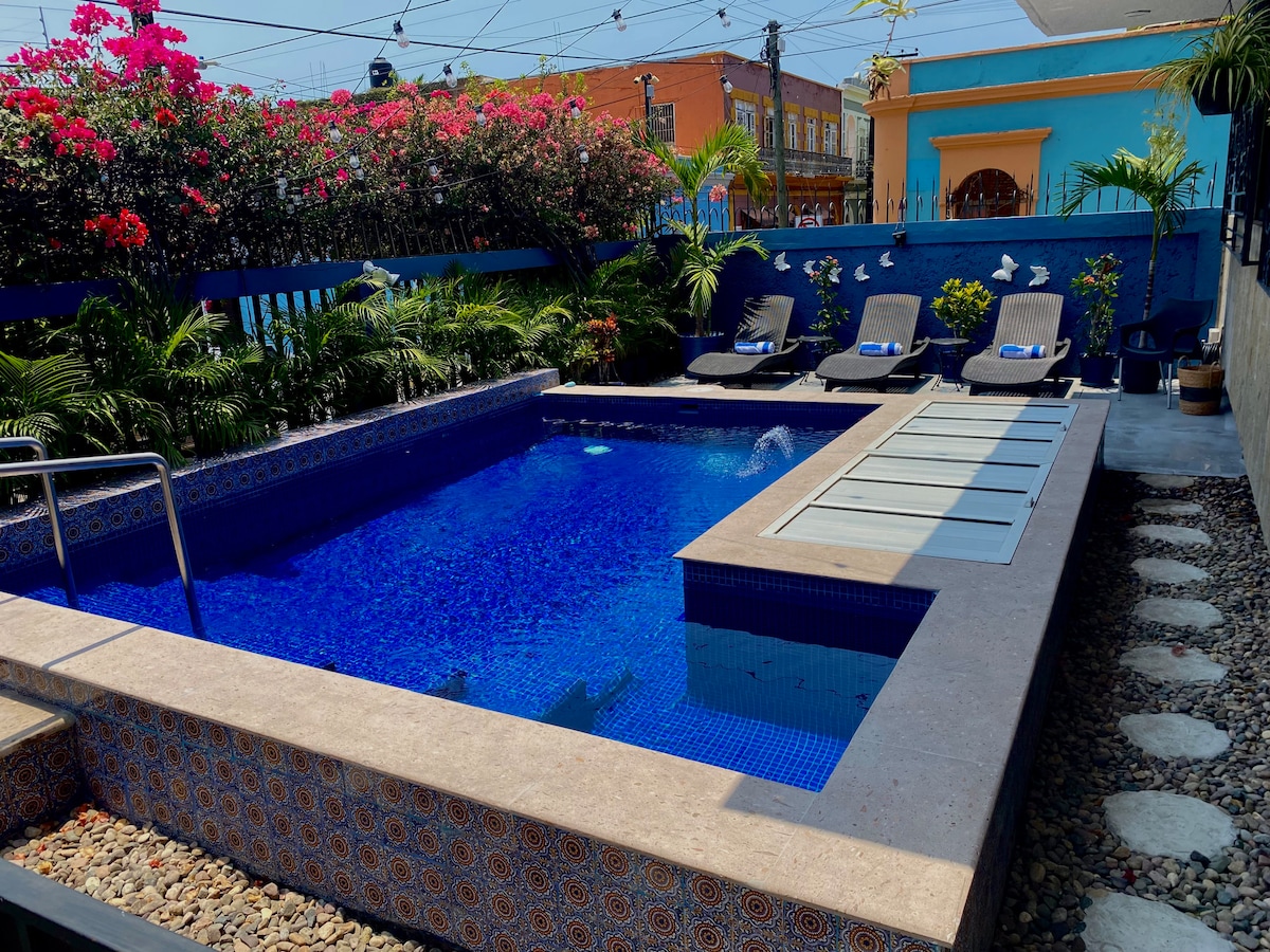 A sparkling blue pool is surrounded by lush greenery and colorful bougainvillea. Comfortable lounge chairs are positioned nearby for relaxation. The pool area features a stone pathway, enhancing accessibility and outdoor enjoyment.
