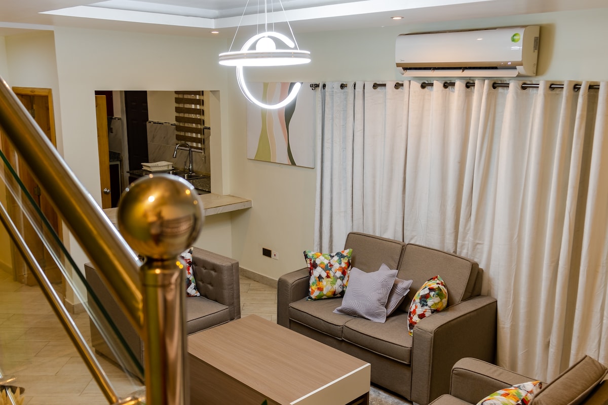 A comfortable seating area features two gray sofas adorned with colorful cushions. A modern light fixture hangs above a wooden coffee table. Light curtains frame a window, while an open area leads to a kitchen space in the background, enhancing the room's functionality.