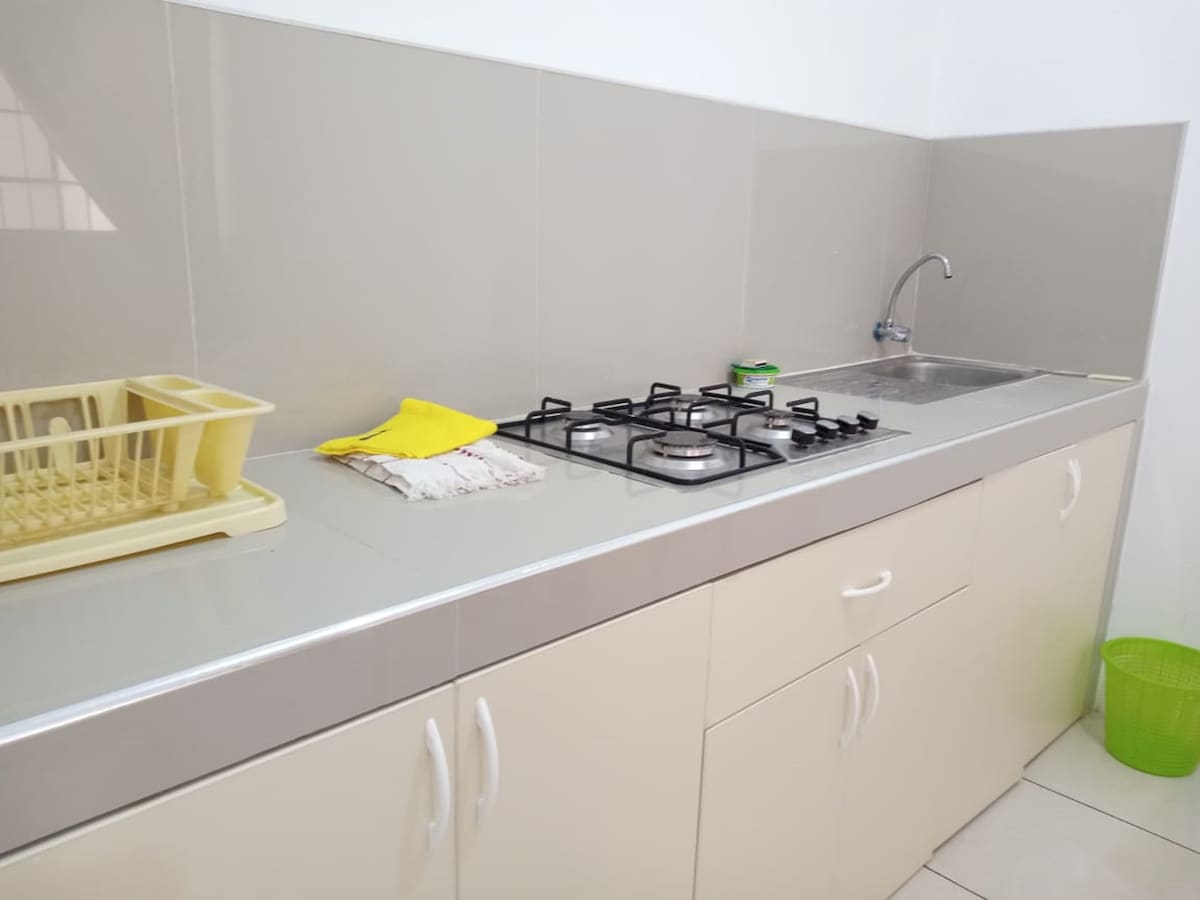 A functional kitchen area is featured with a gas stove and a stainless steel sink. A dish drying rack is positioned beside the sink, and a small green basket is visible on the floor. Neutral-colored cabinets provide ample storage space.