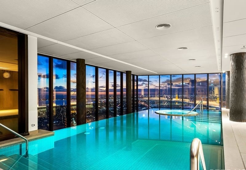 An indoor swimming pool features expansive floor-to-ceiling windows, offering stunning views of the city skyline at sunset. The pool is illuminated in soft blue, with a connected spa area visible. The atmosphere is tranquil, emphasizing the property's upscale design.