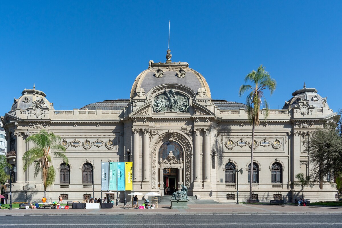 A grand historic building showcases intricate architectural details on its facade. The entrance features ornate sculptures, flanked by two palm trees. Colorful banners are displayed, adding a vibrant touch to the entrance area, which is surrounded by a spacious square.
