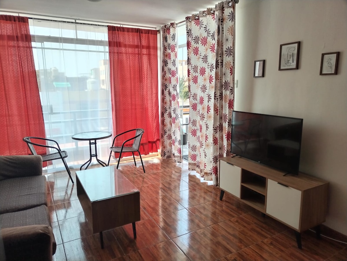 A bright living area features large windows with sheer red curtains and floral-patterned drapes, allowing natural light to illuminate the space. A cozy sofa and a coffee table are positioned in front of a wall-mounted TV. A small dining table with two chairs is near the window.