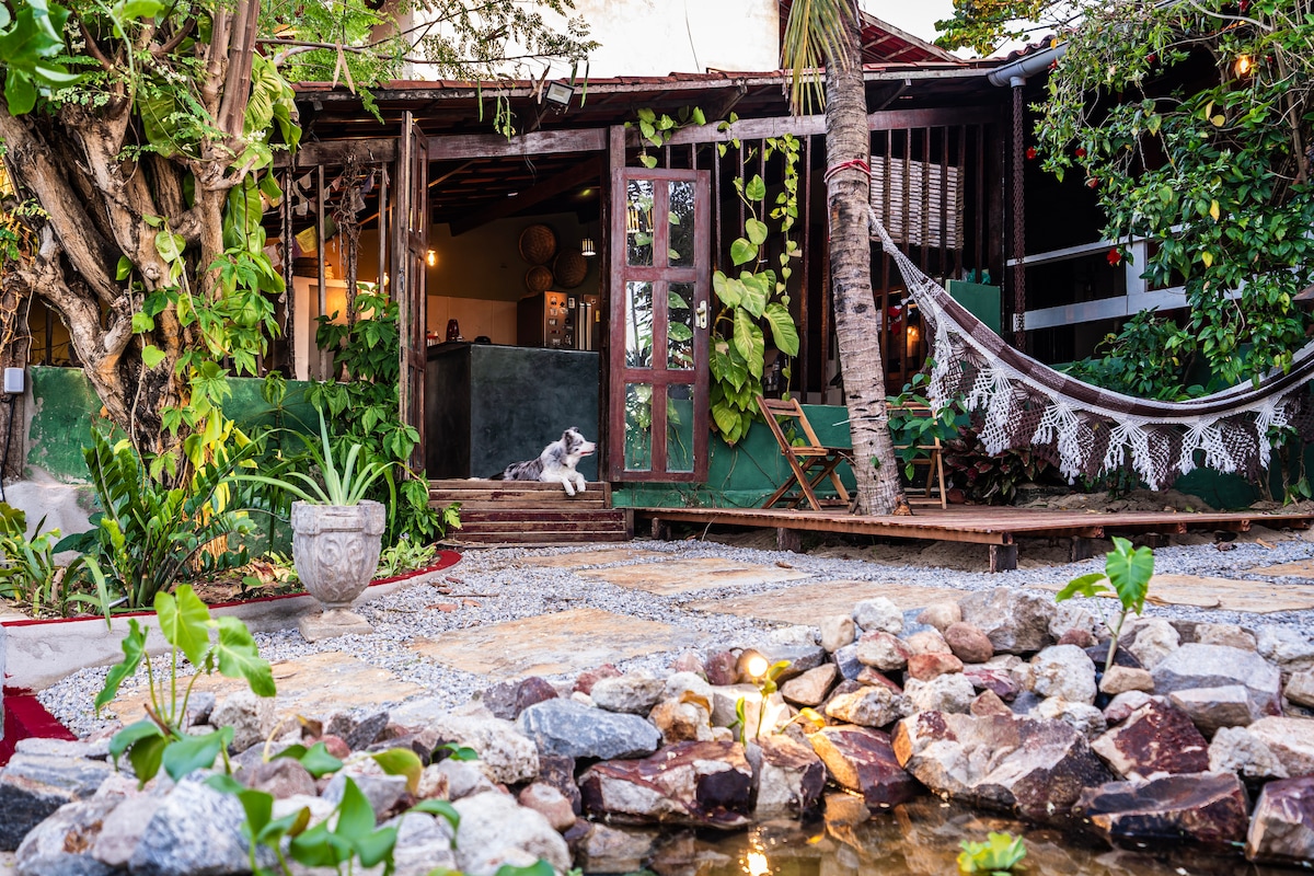 The exterior of the casa is framed by leafy plants and a wooden deck, featuring a hammock for relaxation. A stone pathway leads to an ornate decorative pot. A small pond with pebbles and gentle waterfall adds a calming element to the lush garden.