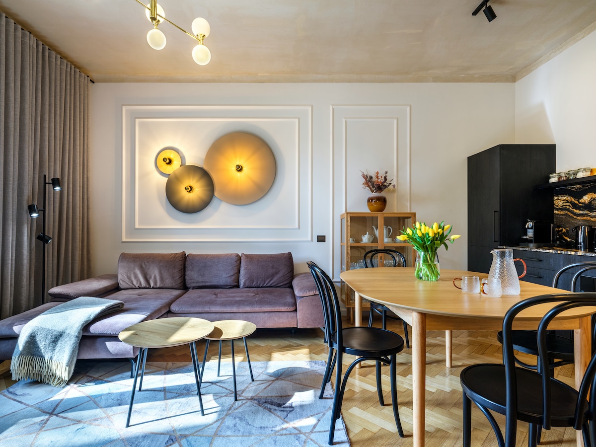 The living area features a spacious sofa accompanied by a set of round coffee tables, enhancing a relaxed setting. A dining table for four is present, complemented by black chairs. Light fixtures and decorative wall art add subtle warmth to the room.