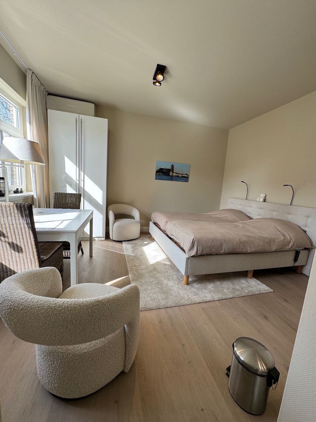 A well-lit sleeping area features a spacious bed with light bedding. Two reading lamps are positioned on either side of the bed. A small desk and two woven chairs are present, along with a cozy accent chair. A wastebasket and a large wardrobe are also visible.