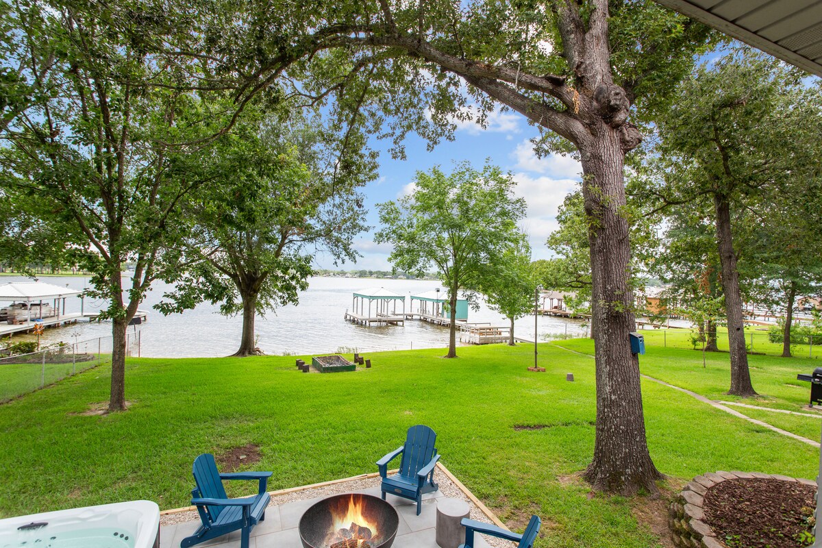 A spacious outdoor area features seating around a fire pit, with a hot tub nearby. Lush green grass surrounds the space, and several trees provide shade. The view extends to a tranquil lake, where a private boat dock is visible in the distance.