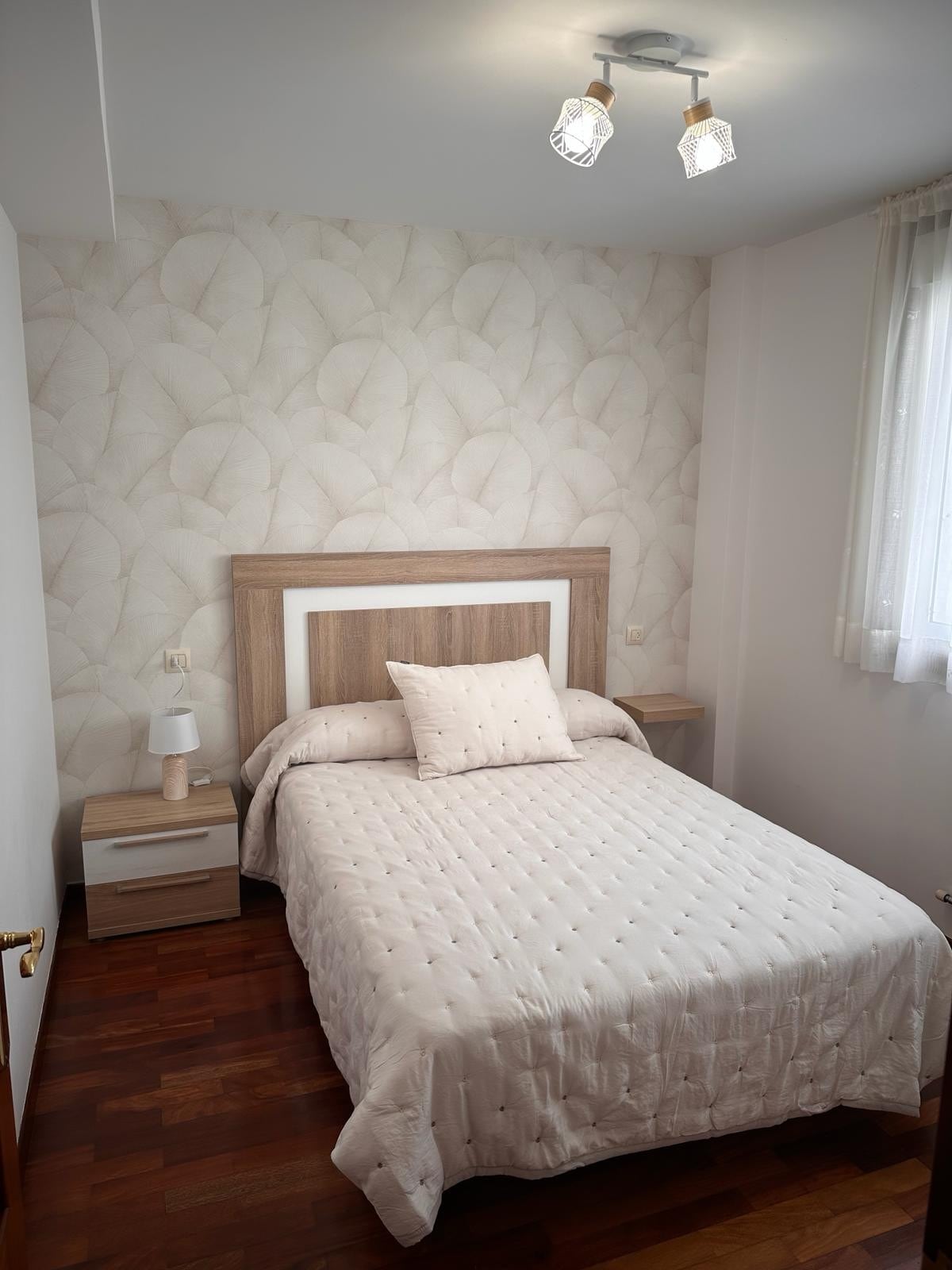A well-lit bedroom features light-colored wallpaper with a subtle leaf pattern. A compact bed is neatly dressed with a quilted coverlet and a decorative pillow. Nightstands are positioned on either side of the bed, while natural light filters in through a window covered by sheer curtains.