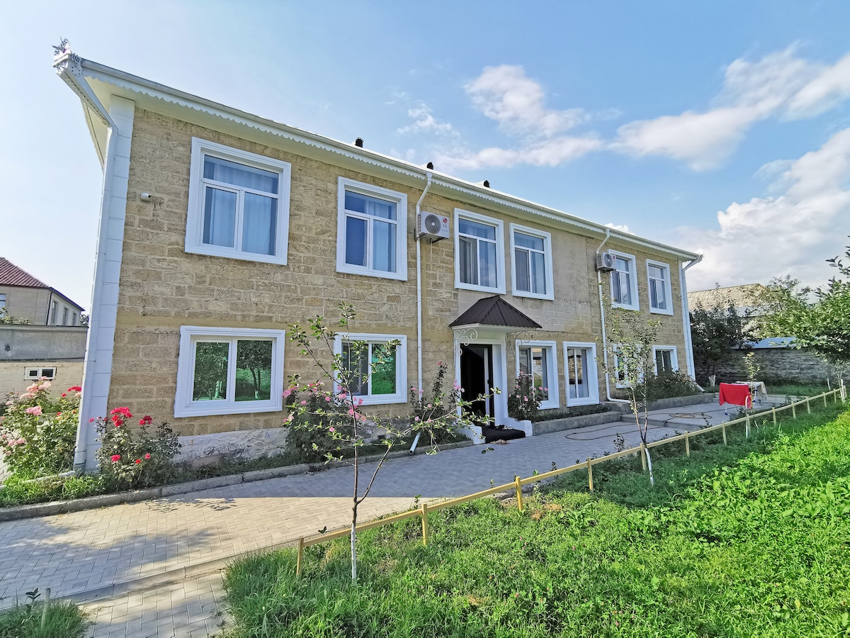 A spacious stone building is set against a clear blue sky. Large windows allow natural light to fill the interior, while a well-maintained pathway leads to the entrance. Lush green grass and blooming rose bushes enhance the outdoor area.