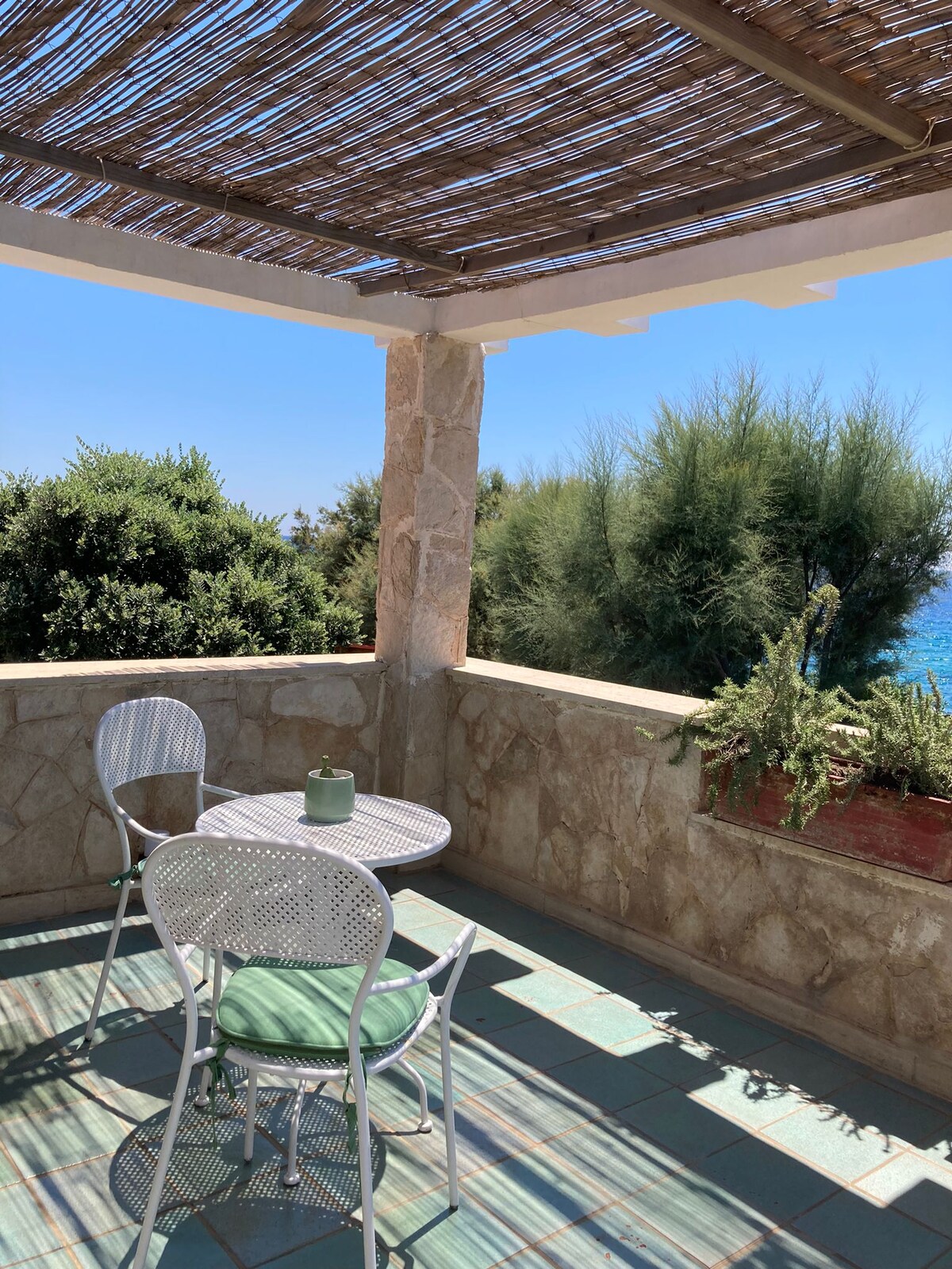 A shaded terrace features a round table with two wire chairs, creating a cozy space for relaxation. Lush greenery is visible in the background, with distant views of the sea shining in the sunlight. Natural materials enhance the peaceful outdoor setting.