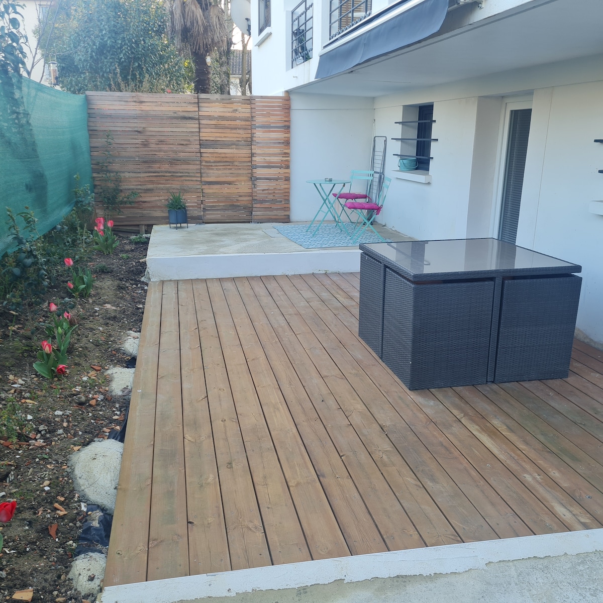 An outdoor terrace features a wooden deck, complemented by a small square table with chairs. Colorful flowers are visible in the garden area, alongside a textured rug. A privacy screen and wall provide separation while enhancing the inviting space for relaxation.