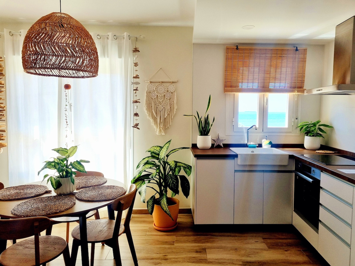 A bright kitchen and dining area features a round wooden table surrounded by four chairs. Lush green plants are placed nearby, and sunlight filters through sheer curtains. A modern kitchen setup includes an oven and a sink, with views of the sea visible through the window.