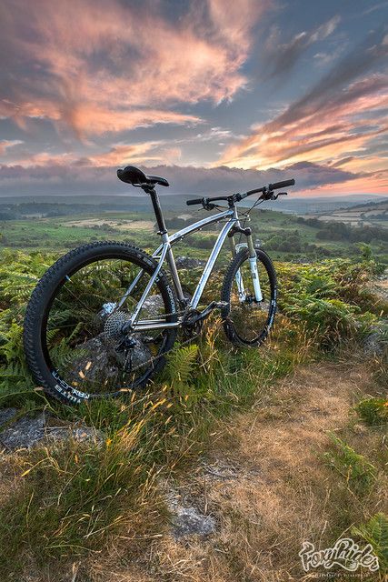 A mountain bike is parked on a grassy hillside, surrounded by fern-like vegetation. The backdrop features a sprawling landscape under a colorful sunset sky, with clouds reflecting shades of orange and purple, creating a serene atmosphere.