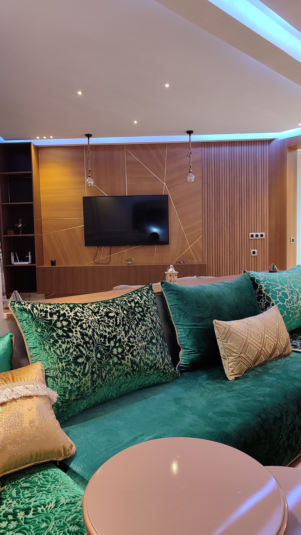 A modern living area showcases a plush green sectional sofa adorned with decorative cushions. A wall-mounted television is positioned above a sleek cabinet. Soft lighting is provided by recessed ceiling fixtures, and a warm wood accent wall adds texture to the space.