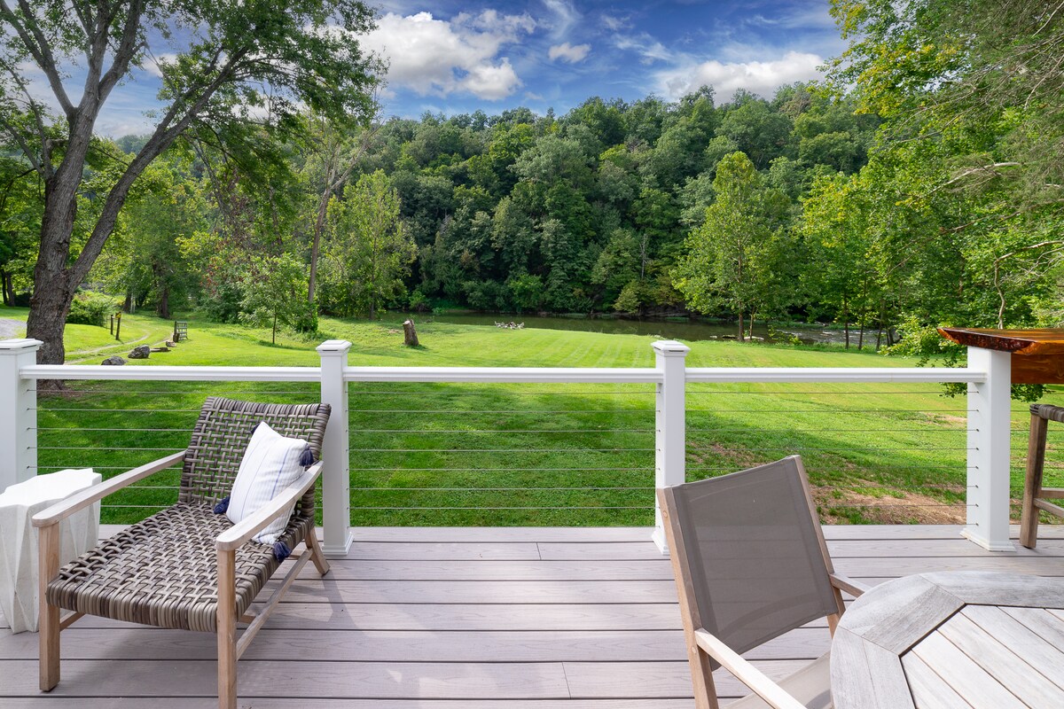 A spacious deck is seen with comfortable seating arrangements, including a woven chair and a wooden table. The scenic view features well-maintained green lawn and trees, with a lush, wooded area in the background under a partly cloudy sky.