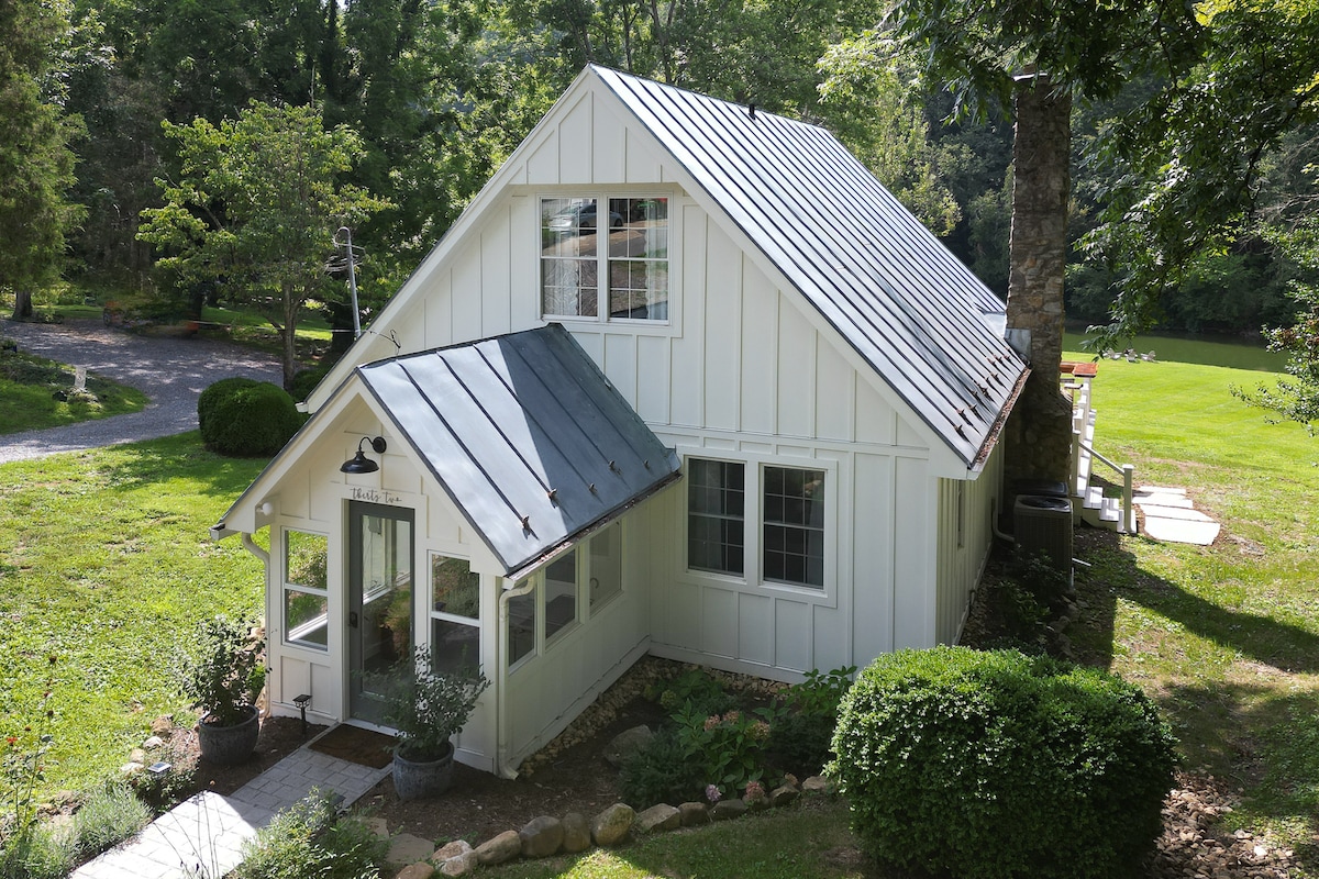 A charming cottage with a white exterior and a metal roof is surrounded by greenery and trees. Large windows allow natural light into the inviting front porch area, which is adorned with potted plants. A gravel pathway leads up to the welcoming entrance.