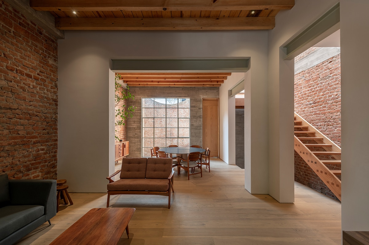 An open living space features exposed brick walls and wooden beams. A cozy seating arrangement includes a sofa and a coffee table. A dining area is visible, surrounded by wooden chairs, with a staircase leading to an upper level illuminated by natural light from large windows.
