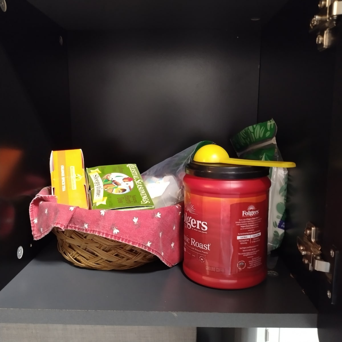A kitchen cabinet houses a woven basket lined with a red fabric, containing assorted items. A canister of coffee sits prominently next to a box of sugar and a bag of green tea, providing essentials for beverage preparation.
