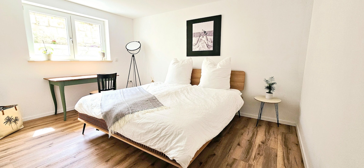 A bright bedroom is presented with a spacious bed featuring two large pillows. A small side table with a decorative plant sits beside the bed, while a desk with a chair is positioned near the window. Natural light filters through the window, enhancing the serene atmosphere.