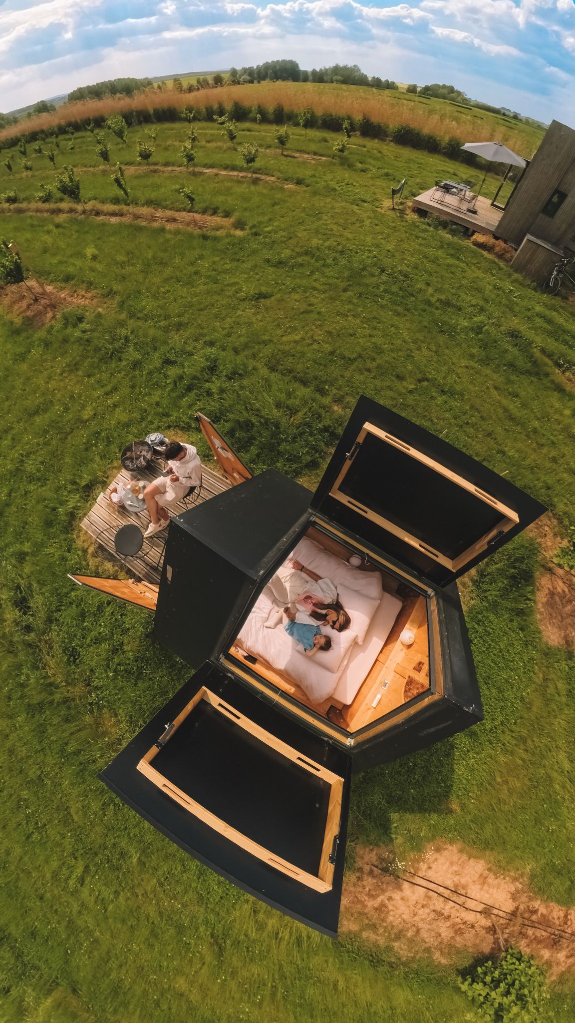 An aerial view captures a tiny house nestled in a green landscape. Open sections reveal a cozy sleeping area. Surrounding fields extend under a blue sky, enhancing the feeling of tranquility. Nearby, additional outdoor spaces are visible, inviting exploration.