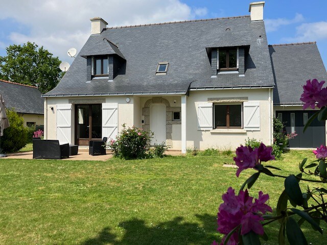 A charming two-story house is set against a blue sky with clouds. The facade features white shutters and a welcoming front door. Lush greenery surrounds the home, and a terrace with outdoor seating can be seen. Colorful flowers add vibrancy to the garden.