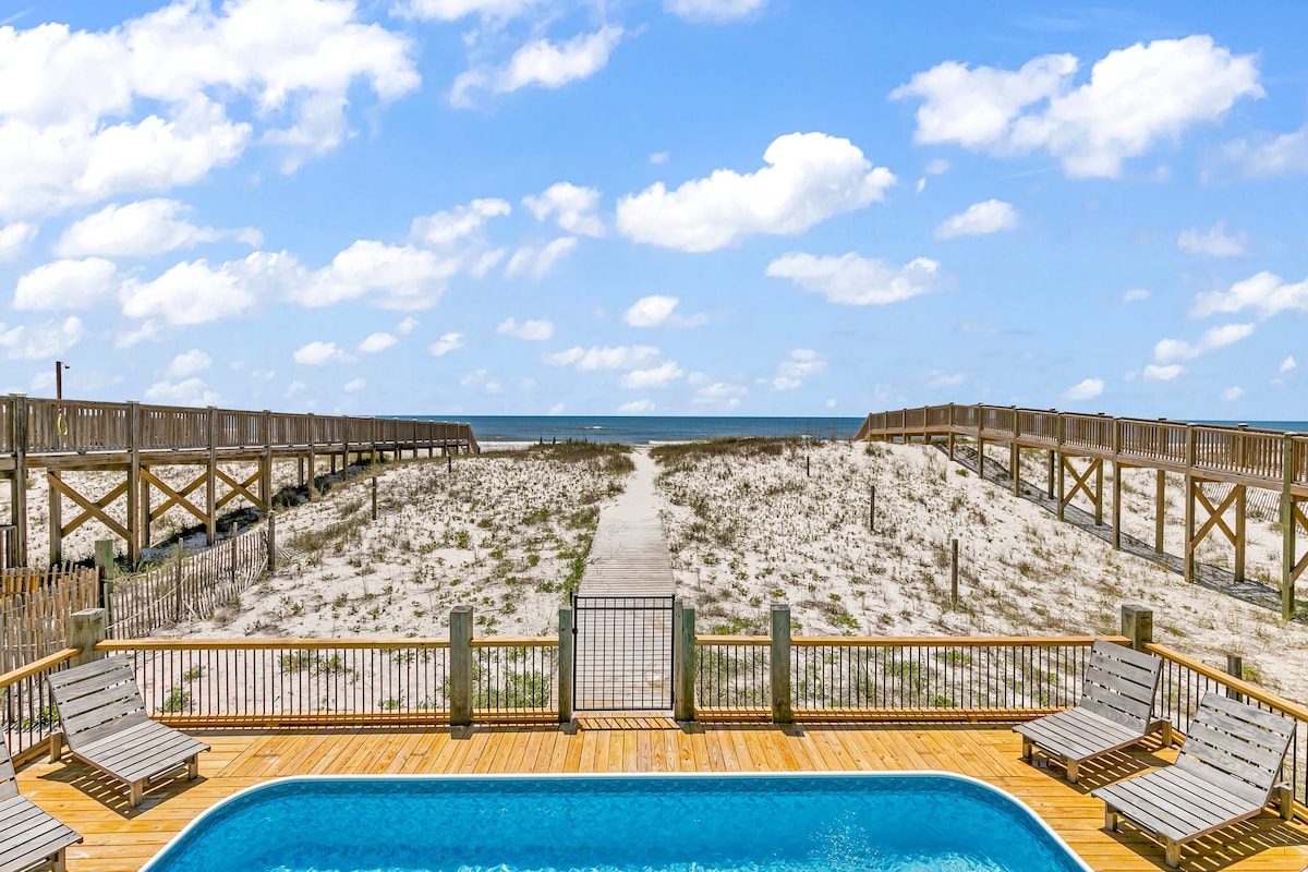 A private pool is visible in the foreground, surrounded by wooden decking with lounge chairs arranged around it. Beyond the pool, a wooden boardwalk leads through sandy terrain towards the Gulf of Mexico, framed by a bright blue sky with fluffy white clouds.