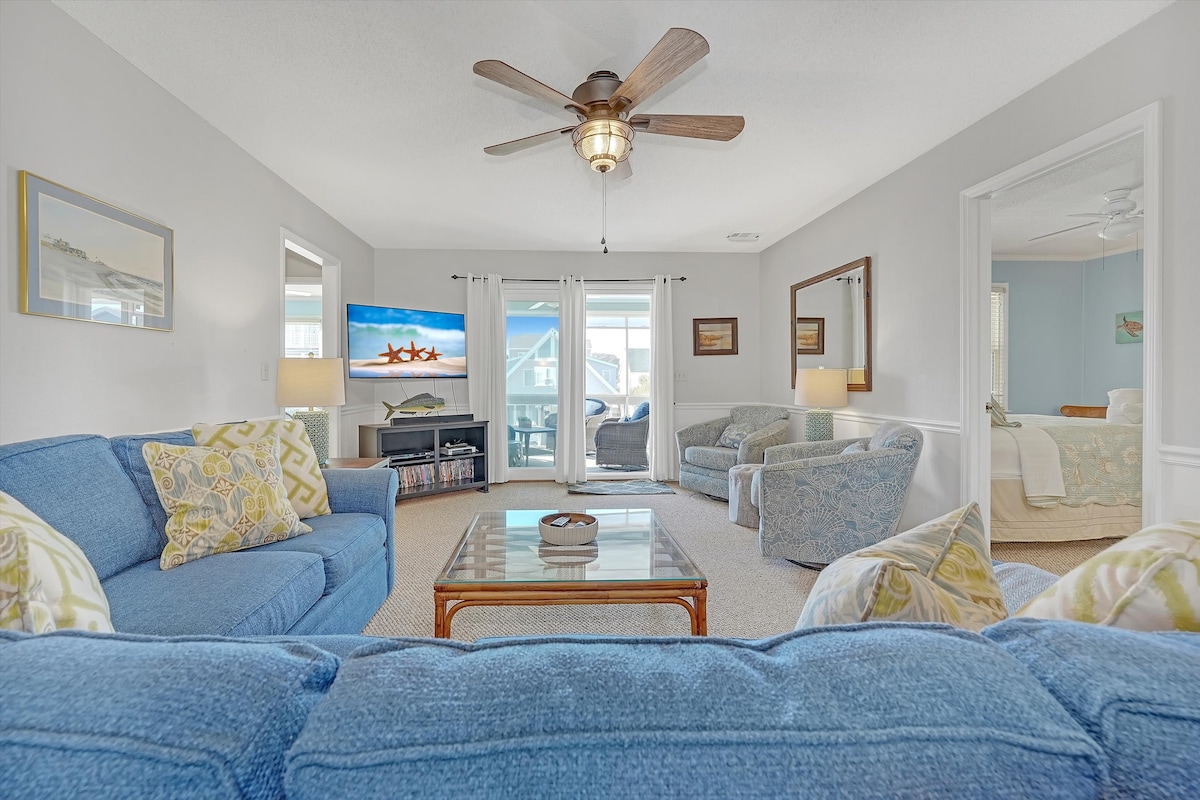 A comfortable living room showcases two blue sofas, accented by colorful throw pillows. A ceiling fan circulates air above, and a coffee table sits at the center. The room is well-lit by natural light coming through large windows, leading to a balcony.