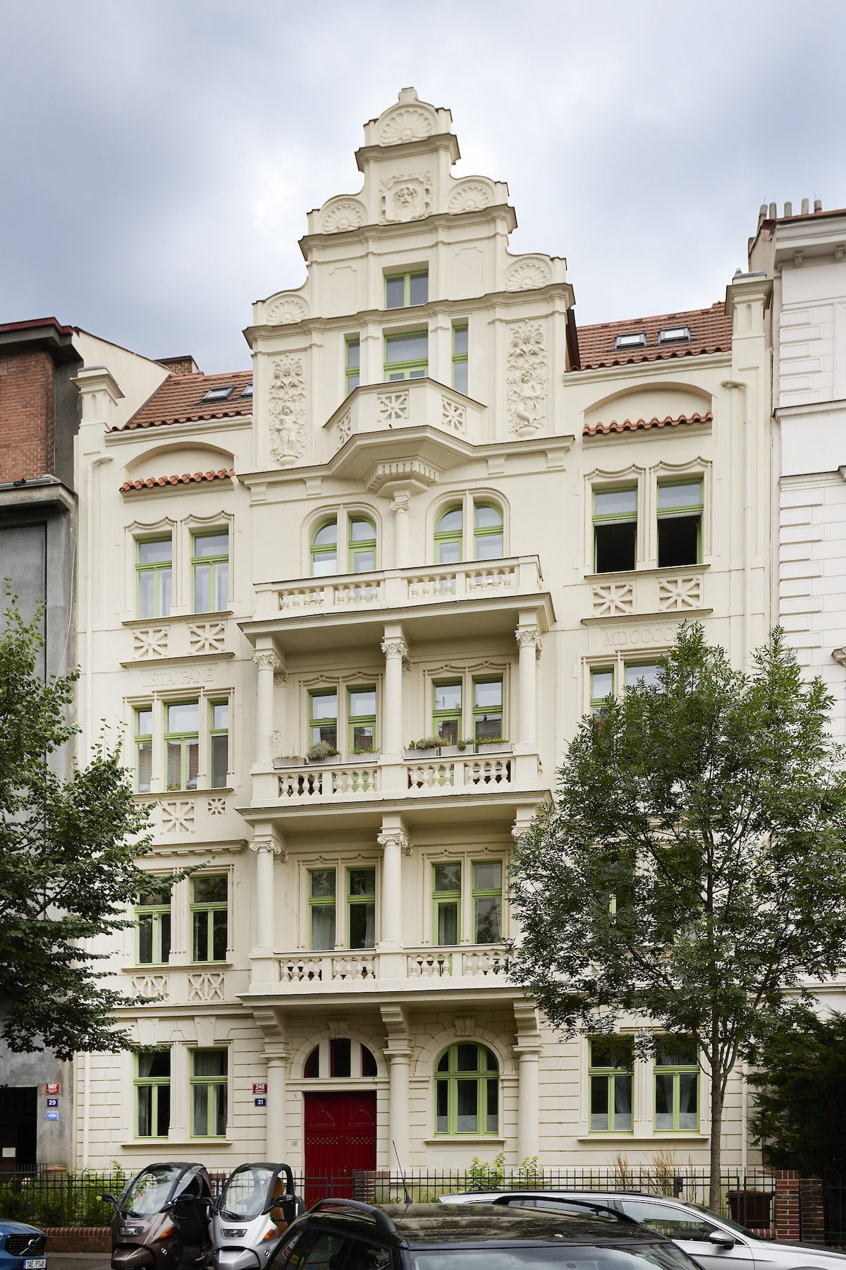 A historic residential building features an ornate facade with intricate architectural details. Numerous windows are symmetrically arranged across the white exterior, accented by green frames. The entrance is marked by a bold red door, flanked by decorative elements and lush greenery at the base.
