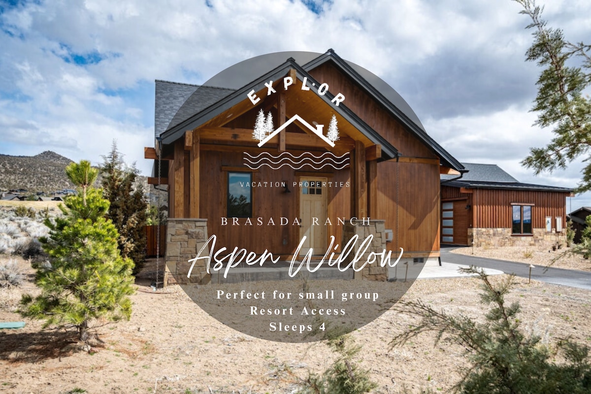 The exterior of a modern two-bedroom home features natural wood siding and stone accents. Surrounded by landscaping, the entrance showcases a welcoming pathway leading to the front door. Trees line the property, contributing to the serene ambiance of the Brasada Ranch Resort community.