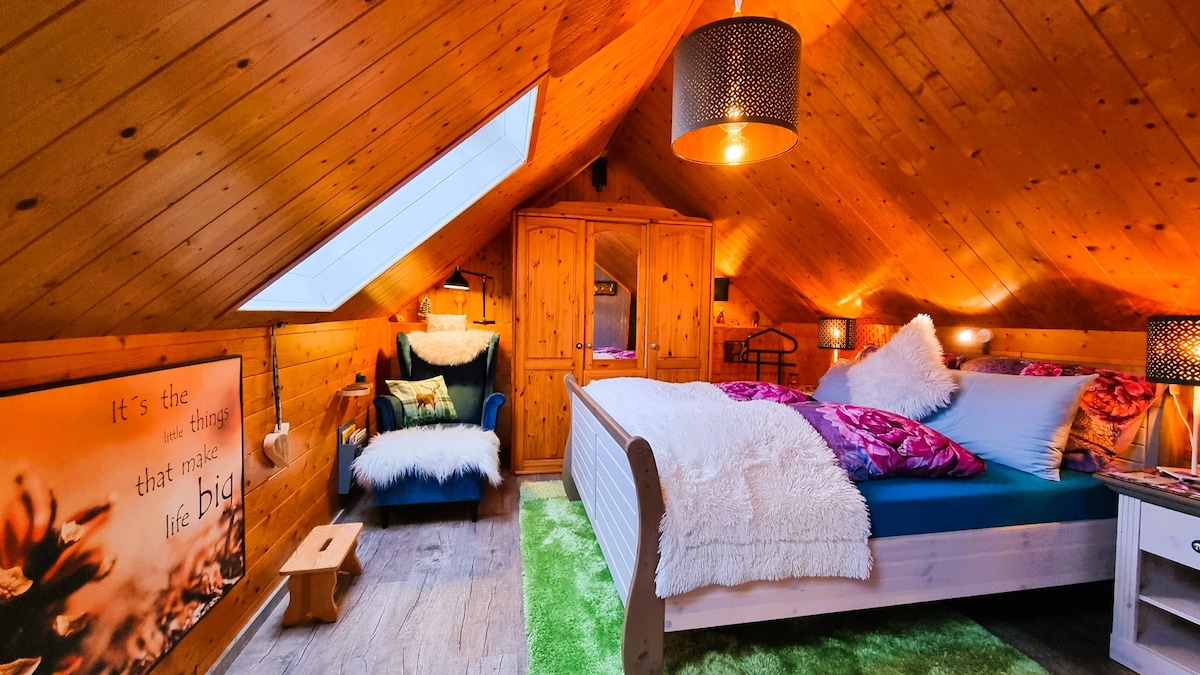 A loft-style apartment features a king size bed adorned with plush pillows and a throw. The warm wood paneling complements a cozy sitting area with a chair and a faux fur throw. A skylight allows natural light to enhance the inviting ambiance of the space.