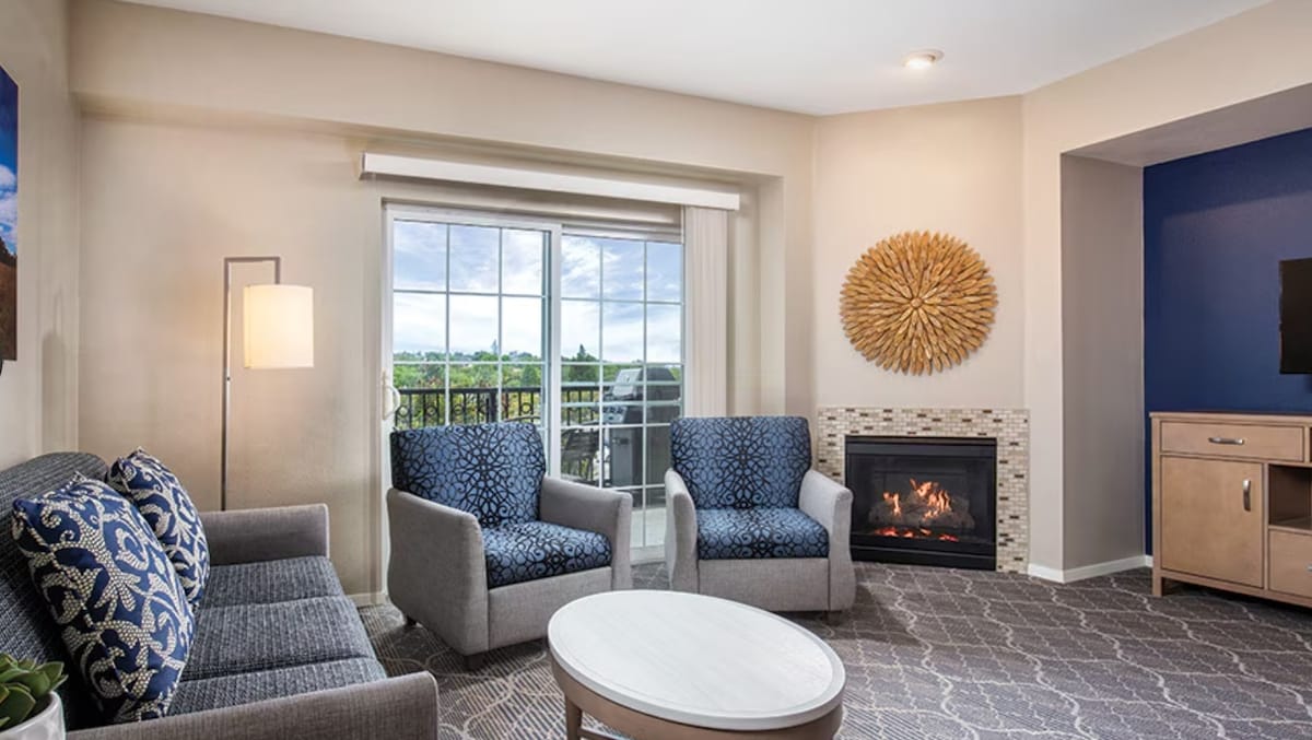 A cozy living area is furnished with two patterned armchairs and a blue sofa, positioned around a round coffee table. A large window allows natural light to brighten the room, while an electric fireplace adds warmth and comfort. Neutral tones create a relaxed ambiance.