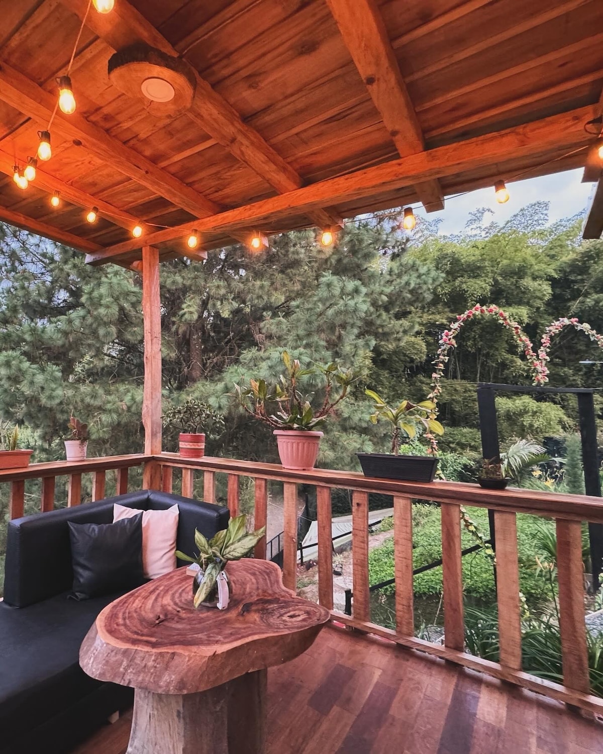 An inviting outdoor patio area is presented with wooden beams and string lights overhead, creating a warm ambiance. A comfortable seating arrangement is visible beside a natural wood table, surrounded by lush greenery and adorned with potted plants, offering a serene escape.