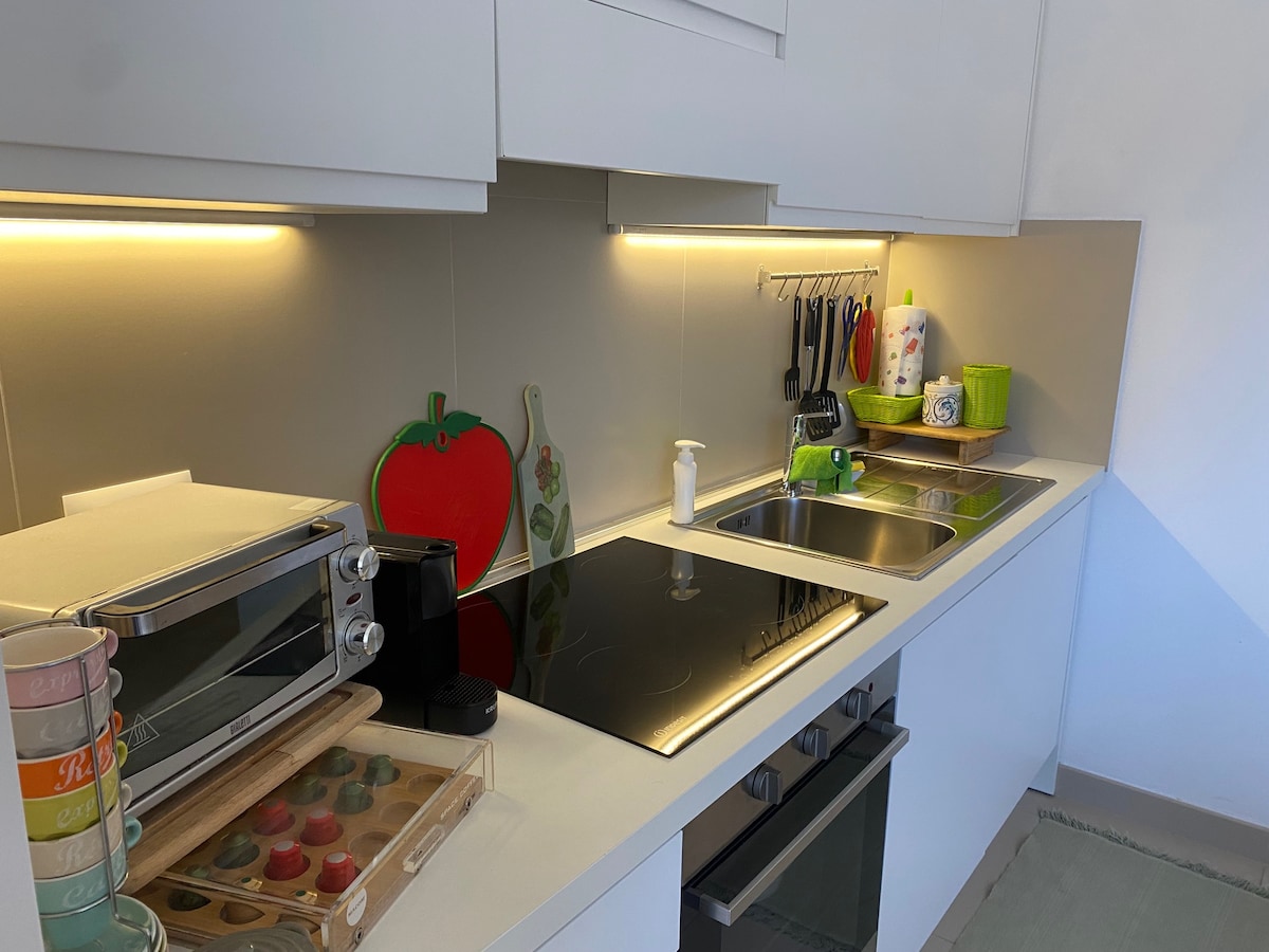 The kitchen features modern appliances, including an oven and a stovetop. Counter space is utilized for various kitchen items, including a coffee maker and a small toaster. Soft lighting enhances the workspace, complemented by colorful decor items on the shelves.