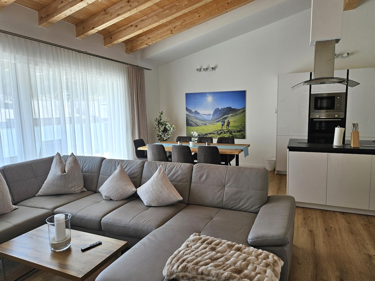 A spacious living area is depicted, featuring a light grey sectional sofa adorned with decorative cushions. A wooden coffee table sits in front, and a large dining table is positioned nearby. Natural light filters through sheer curtains, illuminating the artwork on the wall.