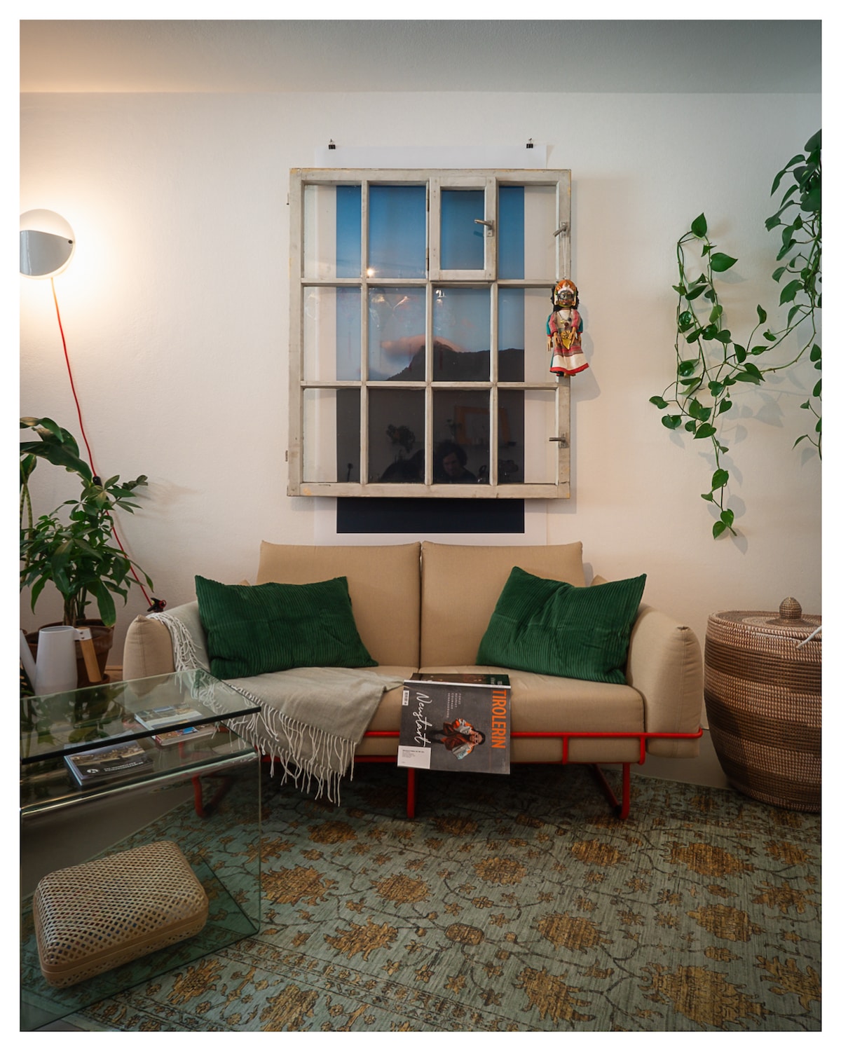 A comfortable sofa is positioned near a window, featuring green cushions for added comfort. A glass coffee table rests in front, adorned with magazines. A potted plant and decorative hanging items bring a touch of nature to the interior, while patterned flooring adds character to the space.