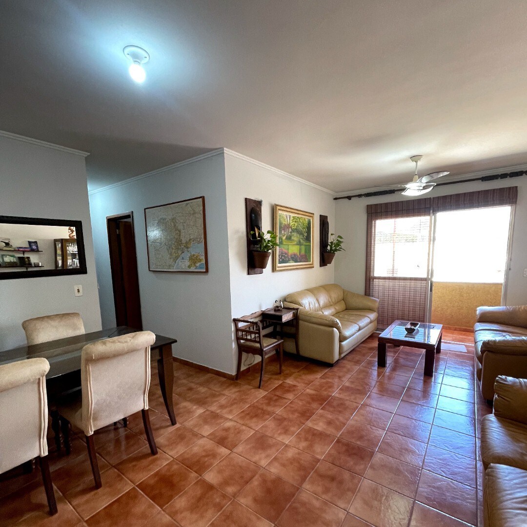 A spacious living area is presented, furnished with two light-colored leather sofas and an elegant coffee table. A dining table with upholstered chairs occupies one corner. Natural light enters through large windows, complemented by partial drapes, while wall art adds a touch of color to the neutral space.